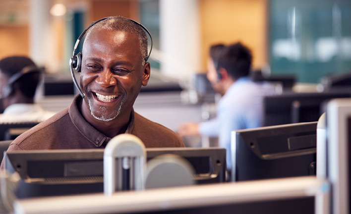 Call center environment with multiple employees working at desks. Foreground features a person wearing a headset, surrounded by computer monitors and office equipment. Faces are blurred for privacy.